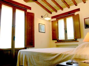 Une chambre à coucher avec un lit, des fenêtres en bois, une lampe fixée au mur et un tableau encadré.