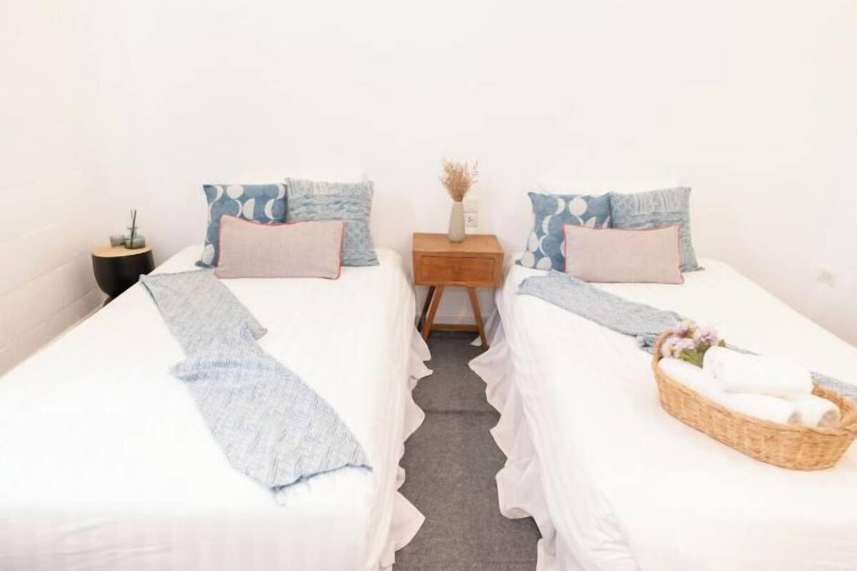 Quarto com duas camas, cada uma com colcha branca e almofadas azuis. Um criado-mudo de madeira com um vaso de flores secas e uma vela. Uma cesta de vime com uma toalha está na cama da direita.