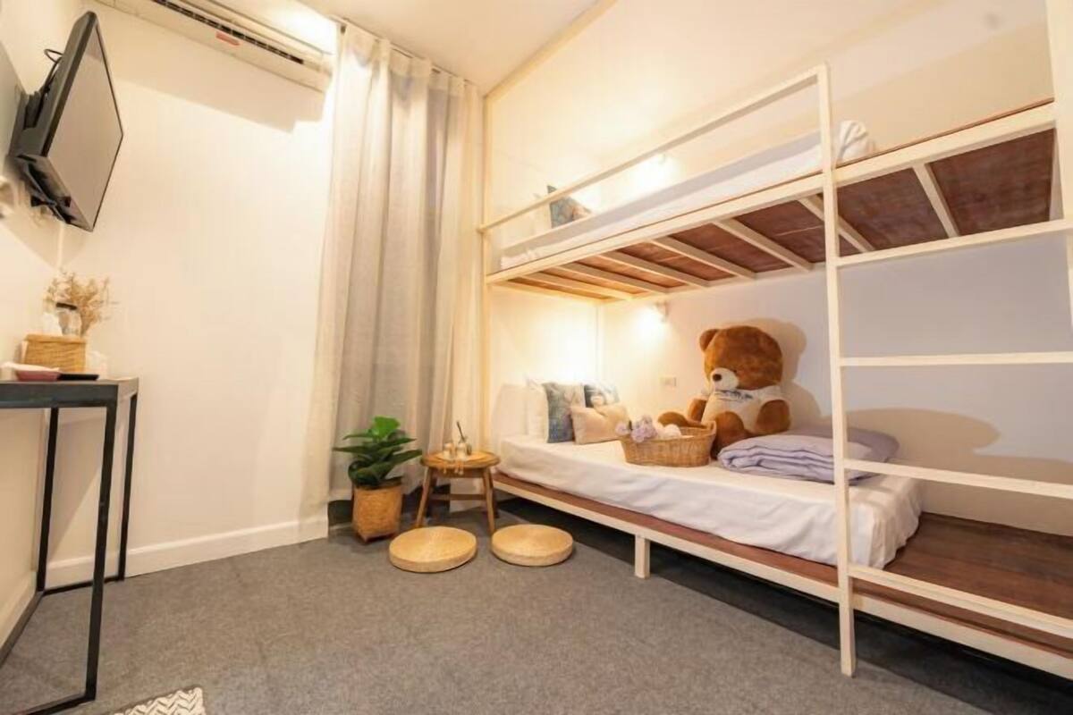 Uma cama beliche com um urso de pelúcia, uma mesinha com uma planta e uma televisão fixada na parede.