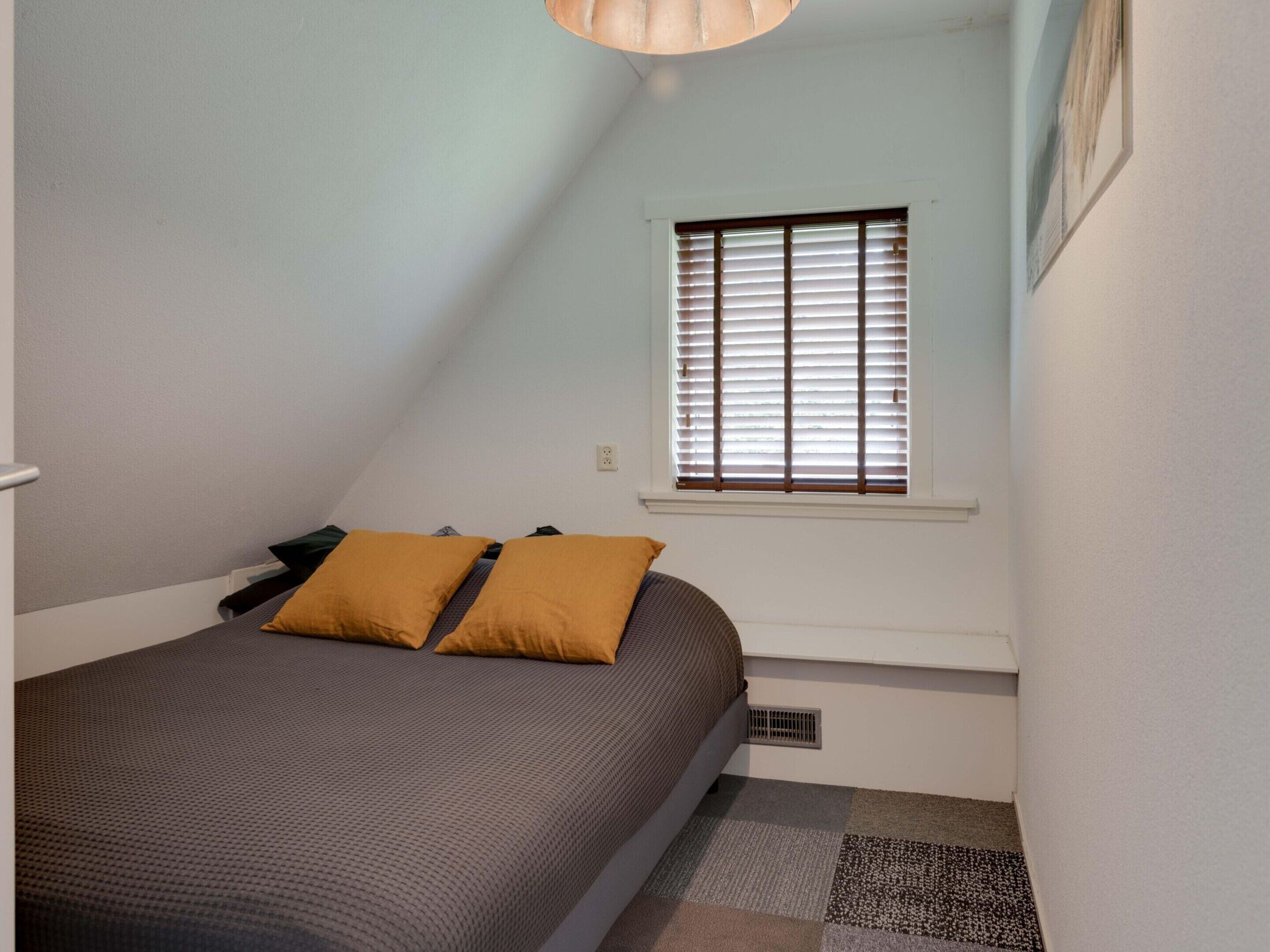 A bedroom with a bed, two pillows, a window with blinds, a light fixture, and a radiator.