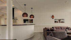 A modern living room with a curved white reception desk, two pendant lights, a grey sofa, and a dining area with a leather bench. - Wellness Villa With Sauna, spa Near Beach (Ouddorp)