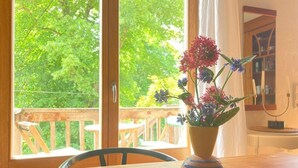 Una mesa de madera con un jarrón a rayas lleno de flores, una silla y vistas a una terraza de madera y follaje al exterior.