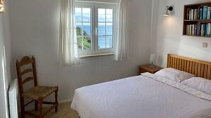 Un dormitorio con una cama grande, una silla de madera, una mesita de noche con lámpara, una ventana con cortinas y una estantería.