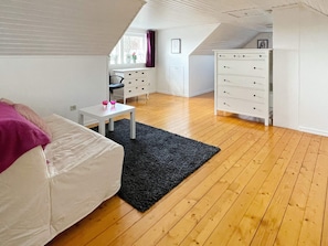 A room with a white sofa, a small table with two pink glasses, a black rug, wooden flooring, a white dresser with drawers, and a window with a curtain. - 4 Star Holiday Home in Farjestaden (Farjestaden)
