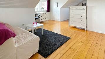 A room with a white sofa, a small table with two pink glasses, a black rug, wooden flooring, a white dresser with drawers, and a window with curtains.