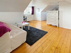A room with a white sofa, a small table with two pink glasses, a black rug, wooden flooring, a white dresser with drawers, and a window with curtains. - 4 Star Holiday Home in Farjestaden (Farjestaden)