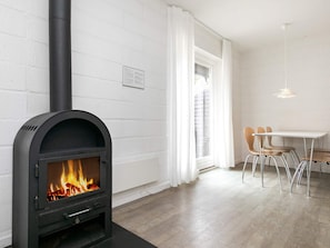 A modern living room with a wood-burning stove, white brick walls, a dining area with a table and chairs, and a window with white curtains. - 6 Person Holiday Park Home in Blavand (Blavand)