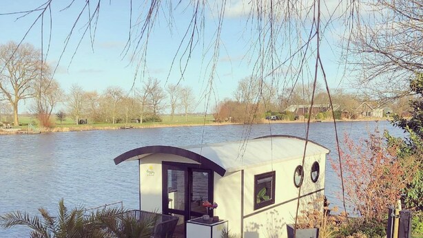 A small houseboat with a deck, windows, and a door, situated by a body of water with trees and a grassy area in the background. - Romantic Boathouse Getaway in Lathum (Lathum)