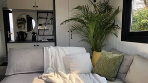 A room with a sofa, a large potted plant, and a built-in shelving unit with kitchen items. - Romantic Boathouse Getaway in Lathum (Lathum)