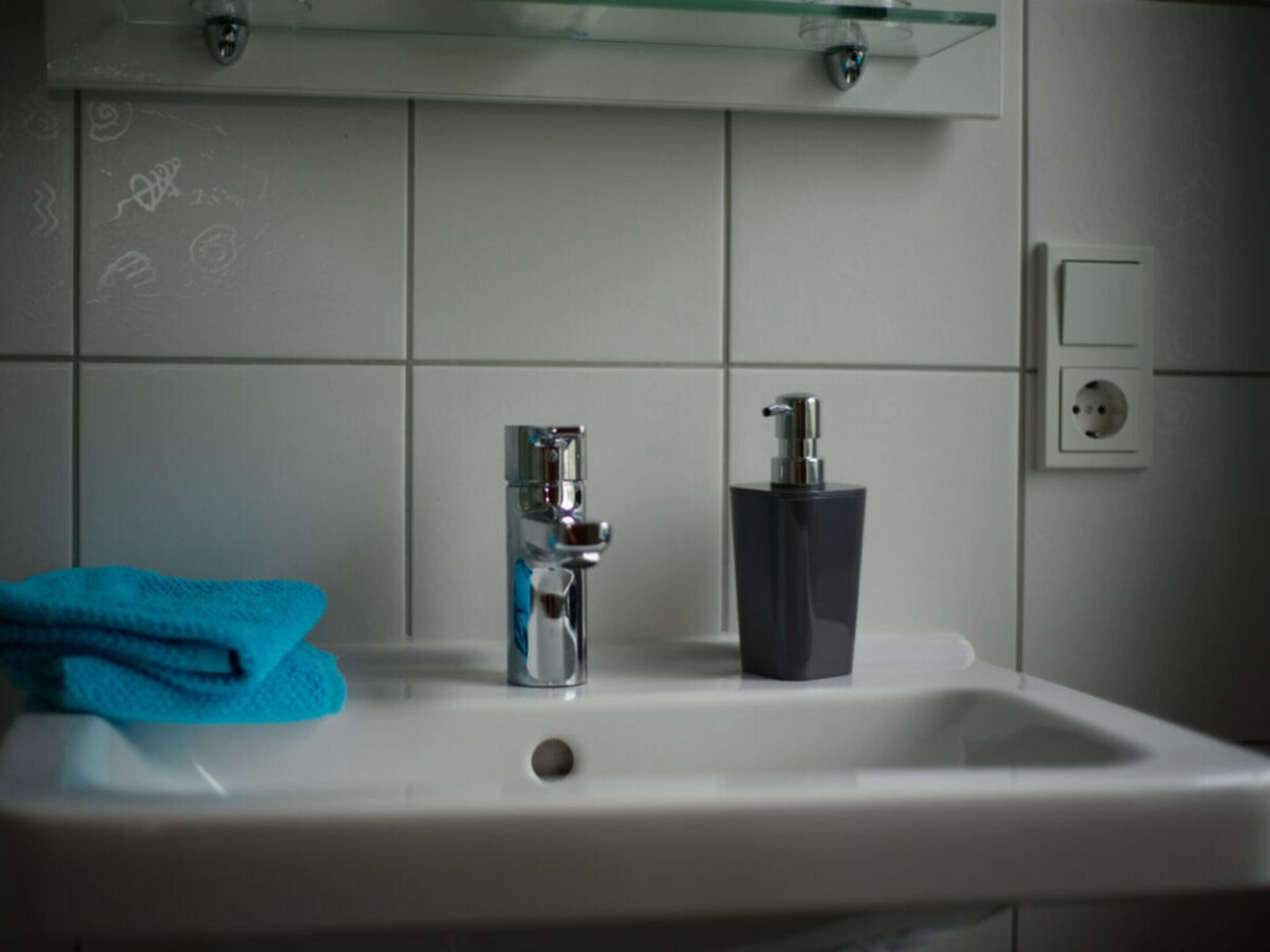 Un lavabo de salle de bain avec un robinet et un distributeur de savon, une serviette bleue et une étagère en verre au-dessus.