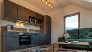 A modern kitchen with dark cabinets, a microwave, and an oven. There's a green sofa with a view of mountains through a large window. - Apartment in Hohentauern Near the ski Area (Hohentauern)