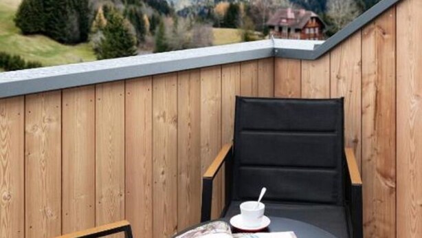 A wooden deck with a black chair and a table, overlooking a mountainous landscape. - Apartment in Hohentauern Near the ski Area (Hohentauern)