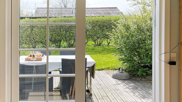 Vista a través de un ventanal a un patio con mesa lista para comer, rodeado de follaje y una terraza de madera.