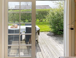 A view through a glass door into a patio area with a table set for dining, surrounded by greenery and a wooden deck. - 6 Person Holiday Home in Struer (Struer)