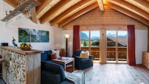 A cozy room with a wooden ceiling, a wooden bar area, a sofa, a chair, and a large window with a mountain view. - No. 2 - Country House Geierstanger (Wallgau)