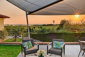 A patio with two chairs, a table with a vase, and a view of a field and sunset.