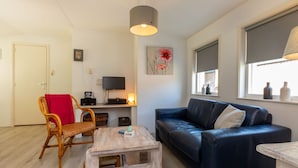 A living room with a dark blue sofa, a wooden coffee table, a wicker chair with a red cushion, a television on a stand, and a painting on the wall. - Cozy Holiday Home 3 Minutes From the Beach! (Zoutelande)