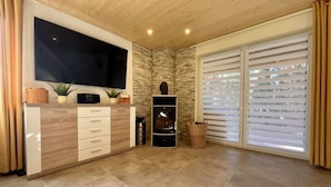 Un salon moderne doté d’un téléviseur à écran plat fixé à un meuble en bois, d’un mur d’accent à l’aspect pierre, d’une cheminée et de grandes fenêtres équipées de stores.