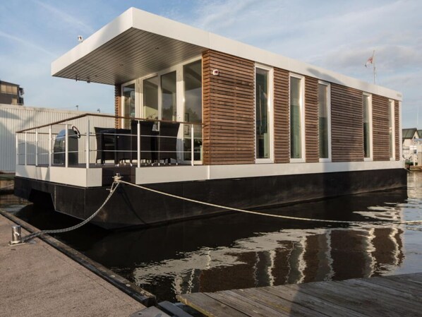 A modern houseboat docked at a pier with a covered deck area and large windows.