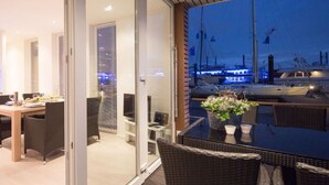 Un balcón con mobiliario de mimbre, una puerta de cristal que da a un comedor y vistas a barcos en un puerto deportivo.