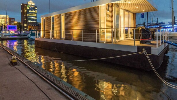 Un edificio moderno frente al agua, con techo plano, ventanas grandes y un espacio exterior cubierto.