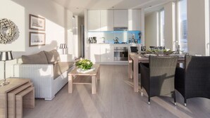 Un apartamento moderno con sala de estar, comedor y cocina. Se observa un sofá, una mesa de comedor con sillas y una cocina con armarios blancos y salpicadero azul.