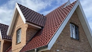 Une maison à deux étages, avec un toit recouvert de tuiles rouges, une façade en briques et un portail vert.