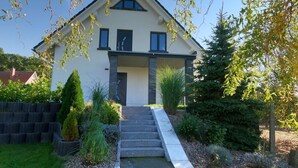 A modern two-story house with a front porch, a small staircase leading to the entrance, and a well-maintained garden with various plants and shrubs. - Golf Idyll Holiday Apartment (Korswandt)