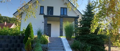 A modern two-story house with a front porch, a small staircase leading to the entrance, and a well-maintained garden with various plants and shrubs.