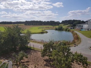 A lakeside view with a building, a paved path, and a fenced area. - Golf Idyll Holiday Apartment (Korswandt)