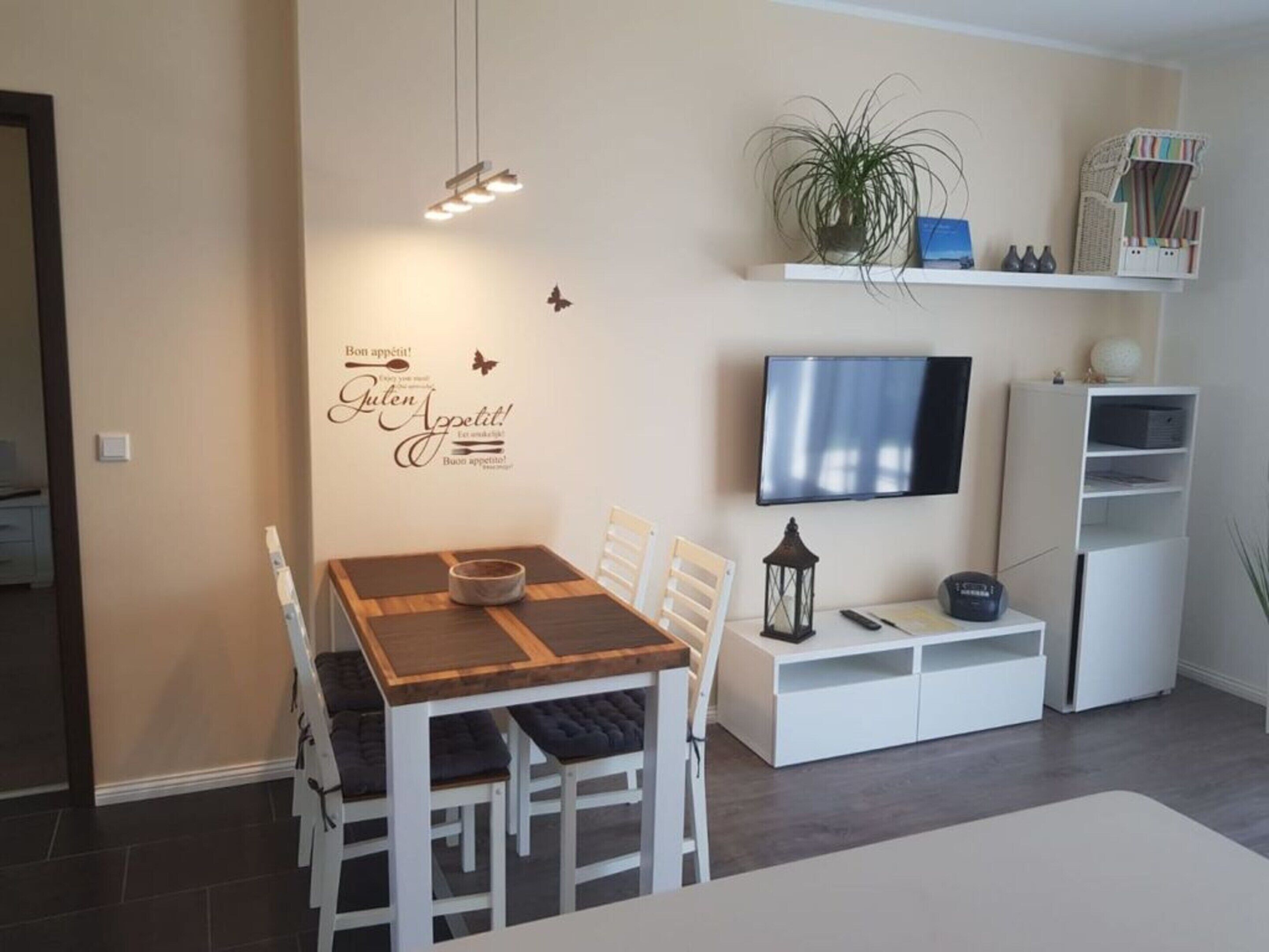 Una sala de estar moderna con una mesa de comedor, sillas blancas, un televisor de pantalla plana, una estantería y una pared decorativa con una planta y una mariposa.