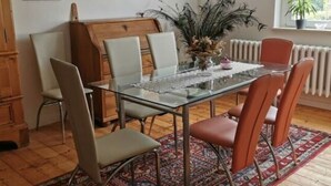 Een eetgedeelte met een glazen tafel, metalen stoelen, een kamerkleed met patroon en een houten kast.