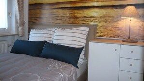 A bedroom with a bed, pillows, a nightstand, a lamp, and a wall mural of a sunset over the ocean. - Holiday Apartment Beach View (St. Peter-Ording)