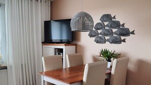 A modern living room with a wooden dining table, white chairs, a flat-screen TV, a decorative fish wall art, and a hanging light fixture. - Holiday Apartment Beach View (St. Peter-Ording)