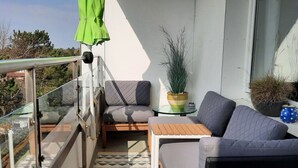 A balcony with a glass railing, a grey sofa, a small table, and a potted plant. - Holiday Apartment Beach View (St. Peter-Ording)