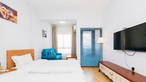 A hotel room with a bed, a TV, a blue chair, a wooden dresser, and a door with blinds. - Hangzhou Tonglu Shengtuo Serviced Apartment (Tonglu East Station Jiangnan Town) (Tonglu)