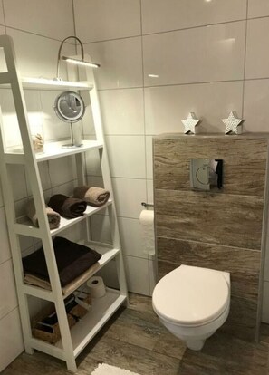 Un baño con inodoro blanco, tocador de madera, repisa con toallas y espejo.