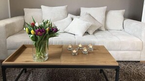 Ein Wohnzimmer mit einem beigen Sofa, weißen Kissen, einer Glaskaraffe mit Blumen und einem Couchtisch aus Holz mit Kerzen.