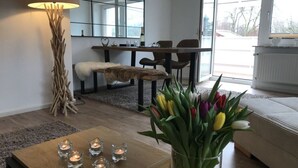 Ein modernes Wohnzimmer mit Couchtisch aus Holz, Kerzen, einer Tulpenvase, einem Fellüberwurf und einem Essbereich mit Stühlen und Tisch.