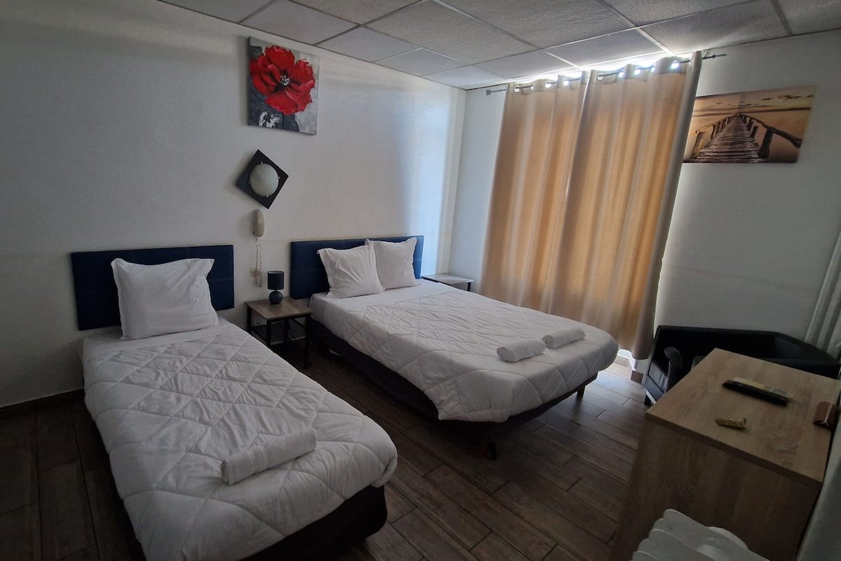 A hotel room with two single beds, a wooden floor, a small table, a lamp, a telephone, and a painting on the wall.