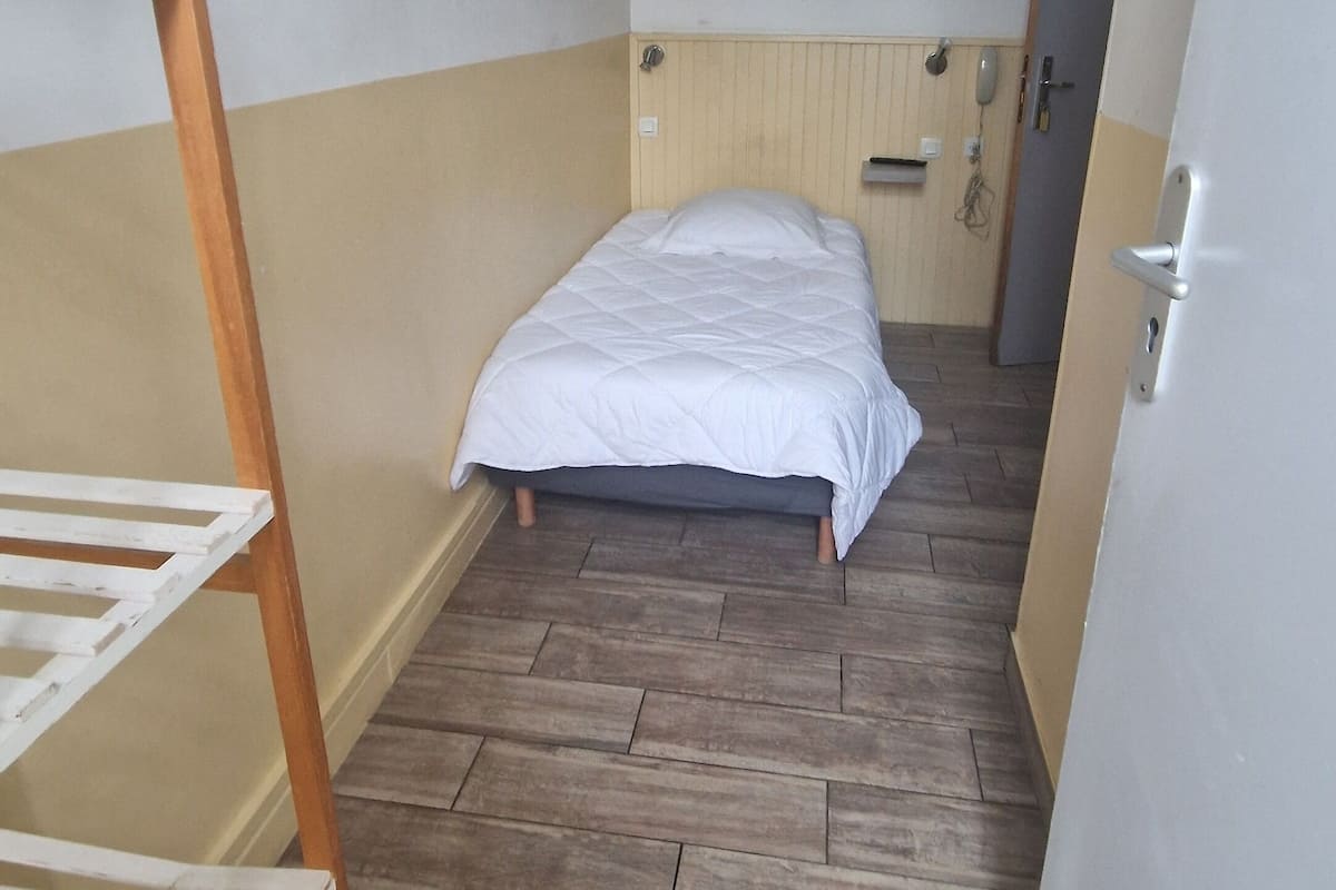A small, narrow room with a bed, wooden floor, and a white door.