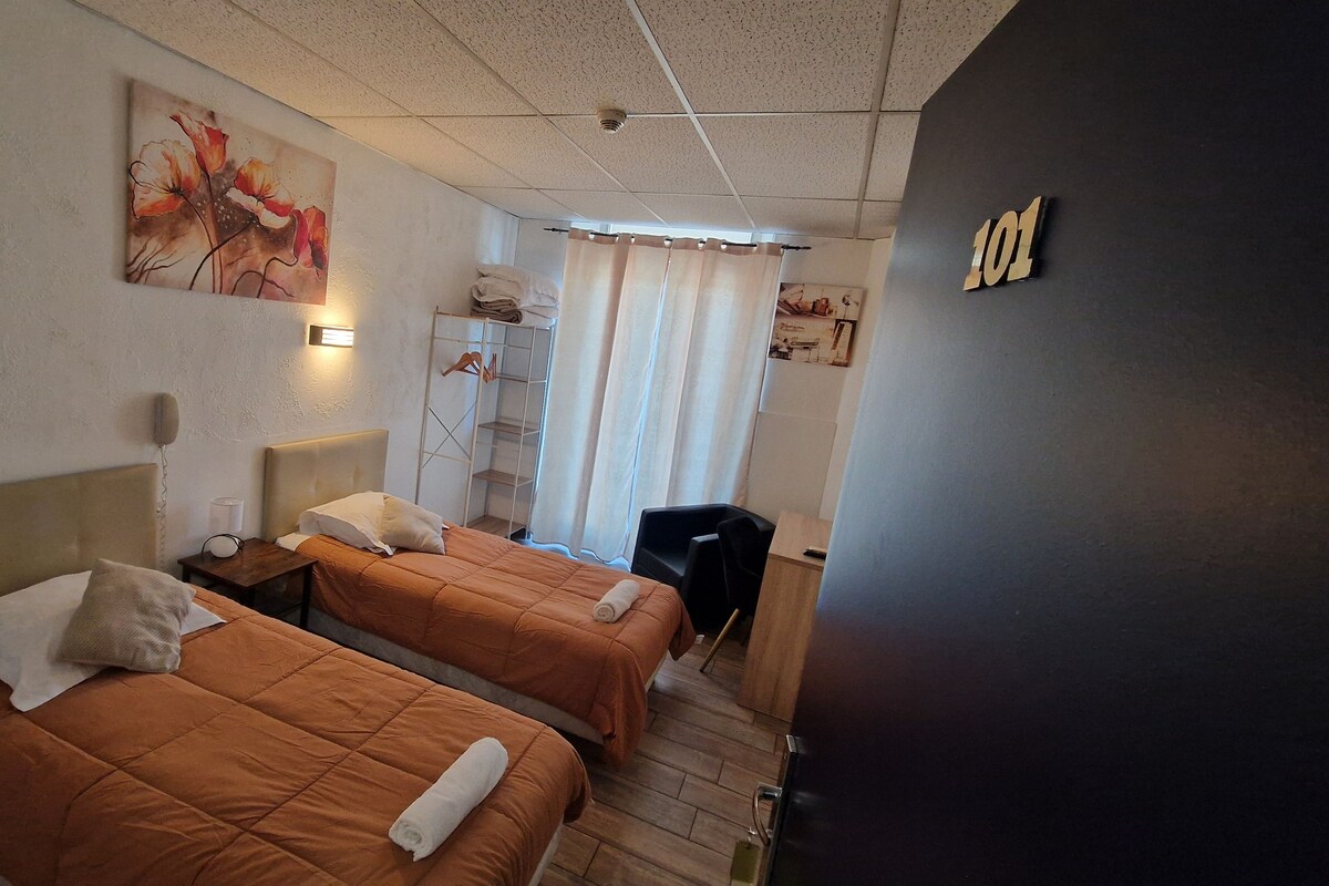 A hotel room with two beds, a bedside table with a lamp, a wardrobe, and a painting on the wall.
