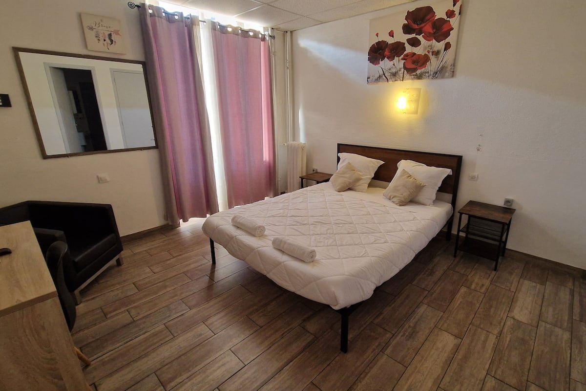 A hotel room with a bed, a desk, a chair, a nightstand, and a wall-mounted light.