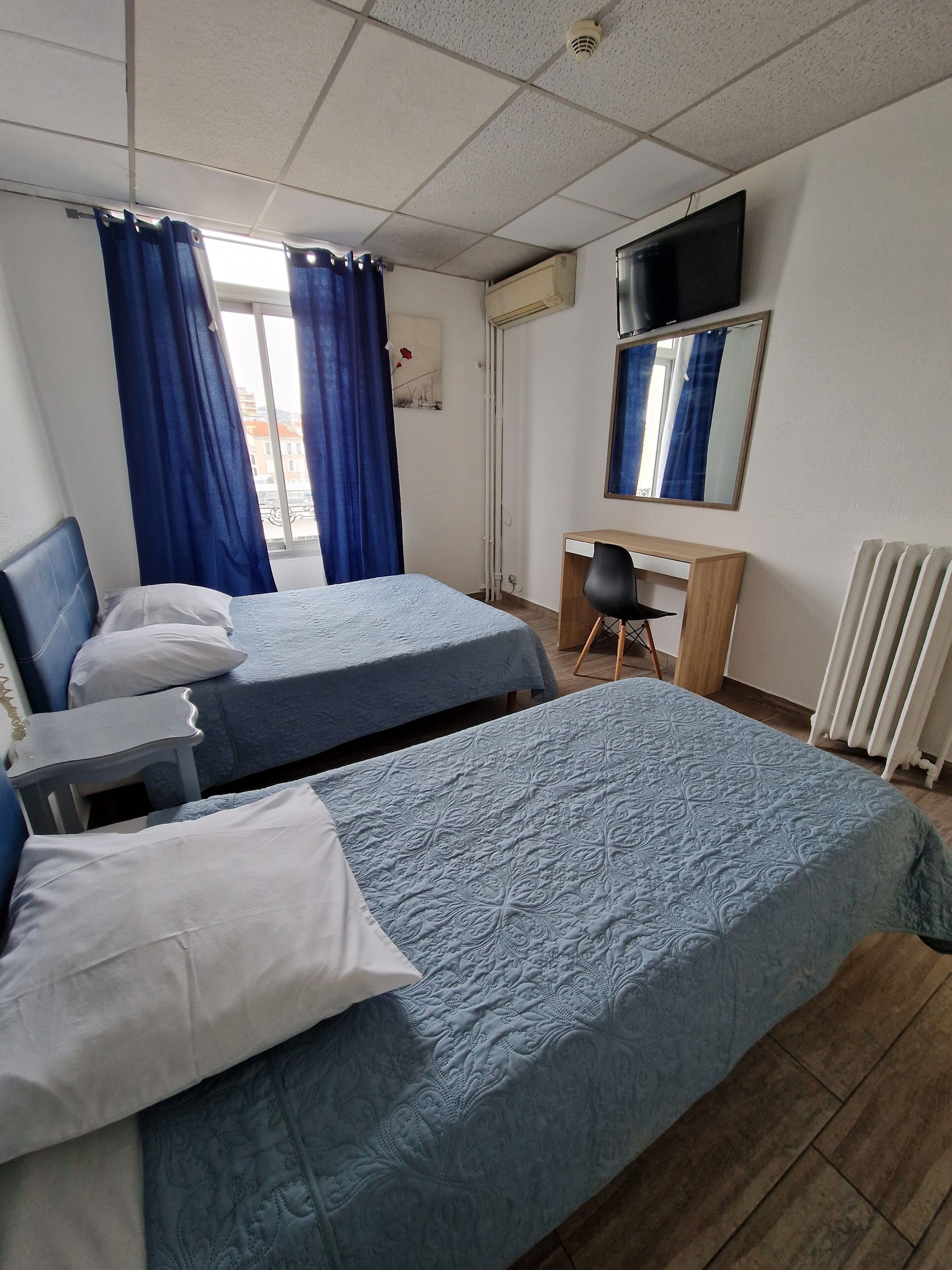 A hotel room with two beds, a desk, a chair, a TV, and a radiator.