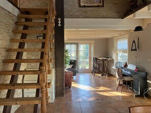 A spacious room with a wooden staircase, a piano, and a dining area with chairs and a table. - Spacious Holiday Home Close to the sea (Burgerbrug)