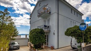 Ein zweigeschossiges Gebäude mit einem Schild, das es als Mehrfamilienhaus kennzeichnet. Vor dem Haus ist ein Auto geparkt, ein kleiner Garten mit Topfpflanzen und der Himmel ist gut sichtbar.