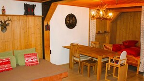 Ein gemütliches Zimmer mit Esstisch aus Holz, Stühlen, einer Couch mit gemusterten Kissen, einem Kamin und einer Holzbalkendecke.