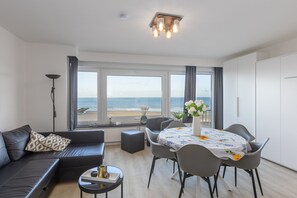 A modern living room with a sofa, dining table, and chairs, large windows with a view of the beach, and a ceiling light fixture. - Cosy Studio With Sleeping Area and sea View (Middelkerke)