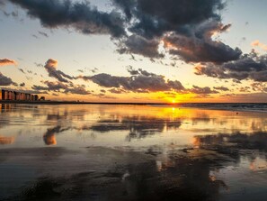 Una playa con un atardecer, agua tranquila y un horizonte urbano distante.