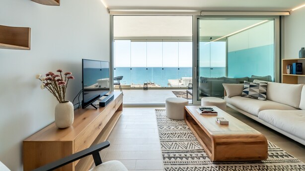 Una sala de estar moderna con un televisor grande, muebles de madera, un sofá y vistas al mar a través de amplios ventanales.
